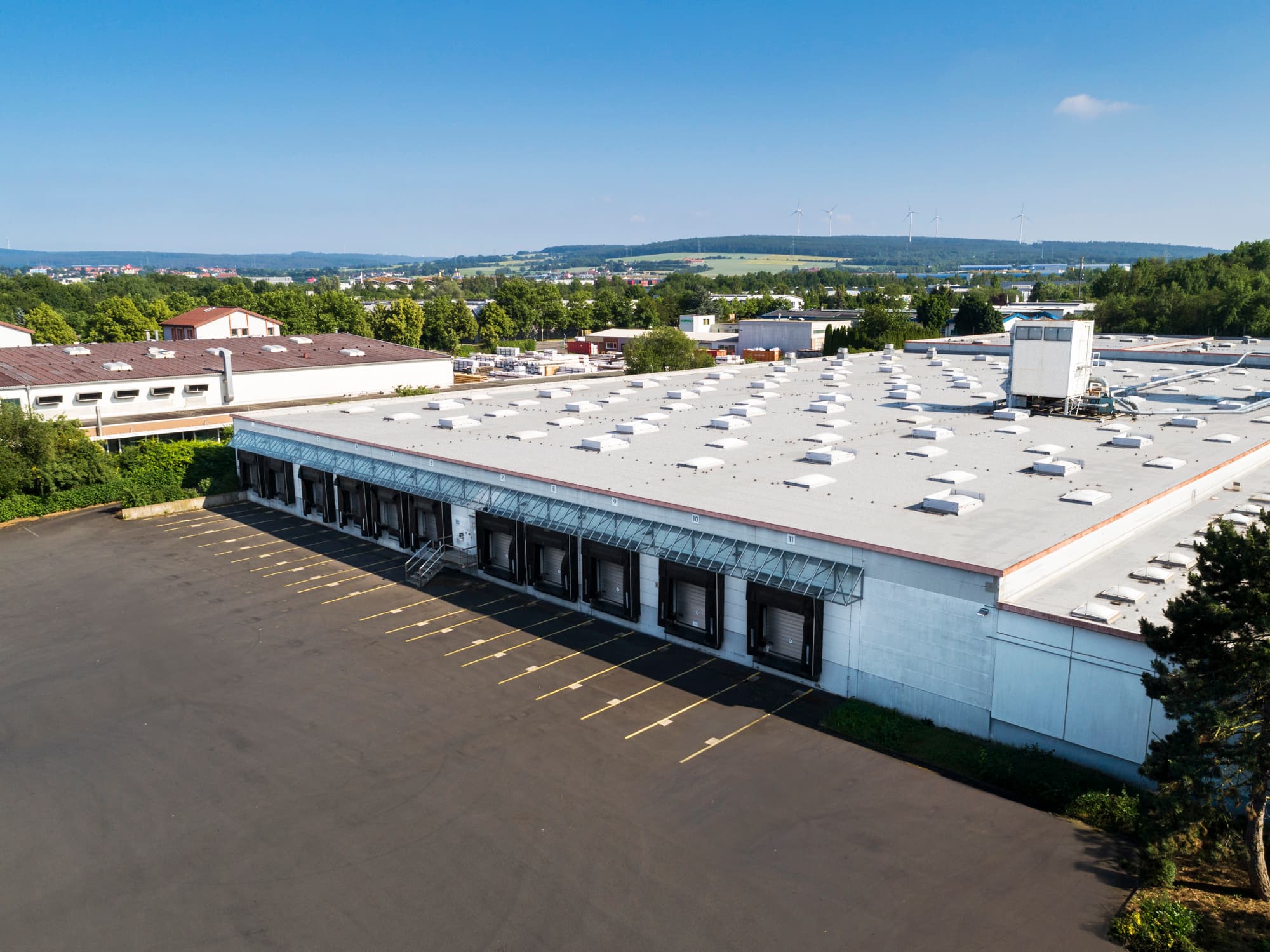 A large industrial warehouse with a white roof and multiple loading dock doors, situated in a paved lot. Surrounding the building is green foliage, and a distant landscape features hills and wind turbines under a clear blue sky.