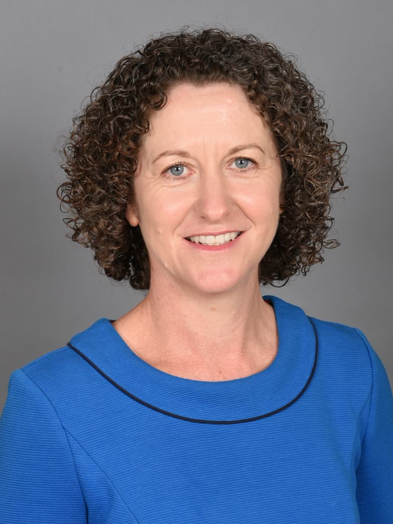 A person with curly hair is smiling and wearing a blue top with a black trim. The background is a neutral gray.