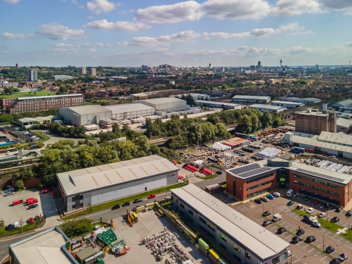 Aerial view of an industrial area with several warehouses, other buildings, and parking lots. The surrounding landscape features green trees and a city skyline in the background under a partly cloudy sky.