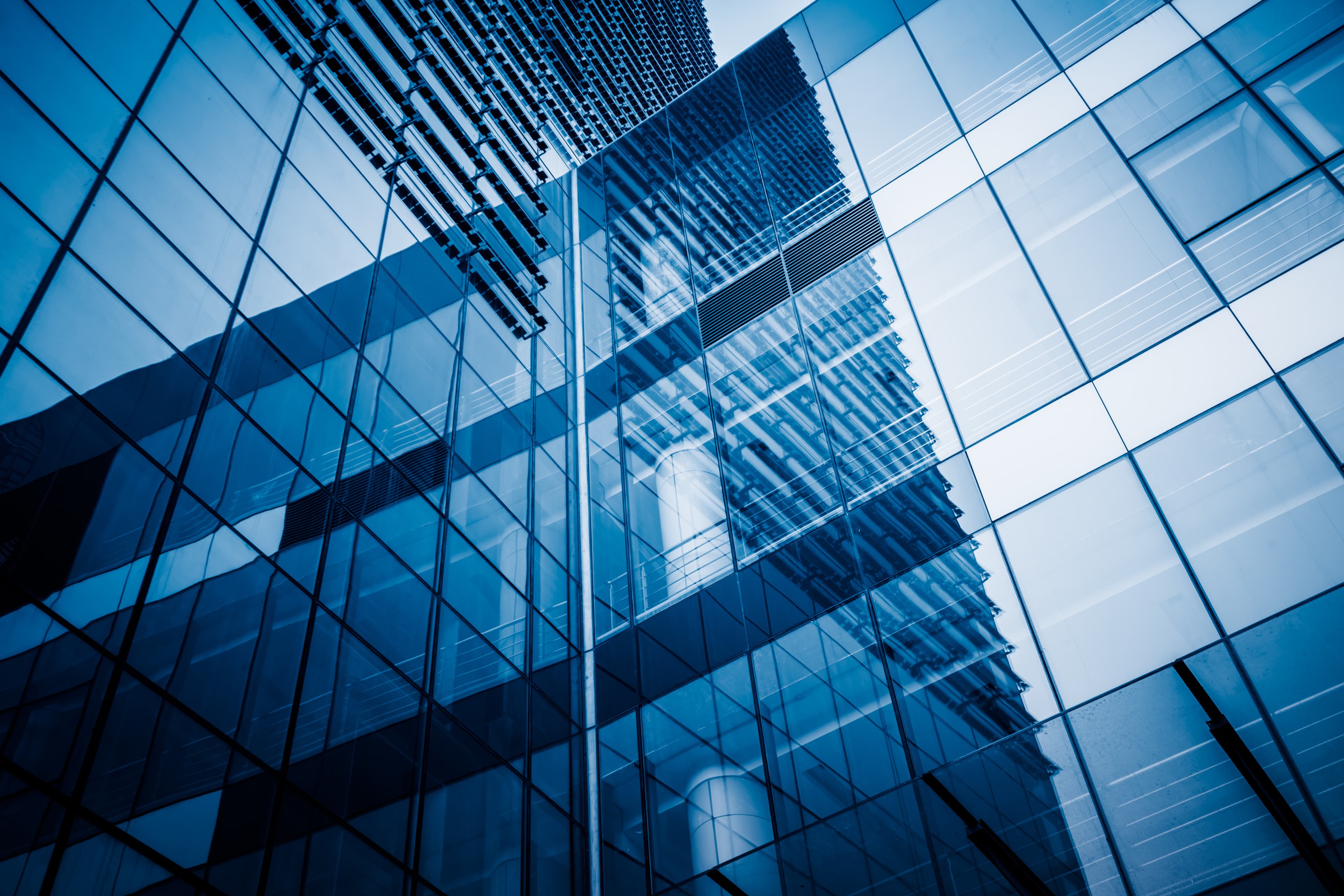 Modern glass building exterior with reflective windows showing the structure and surrounding architecture in shades of blue.