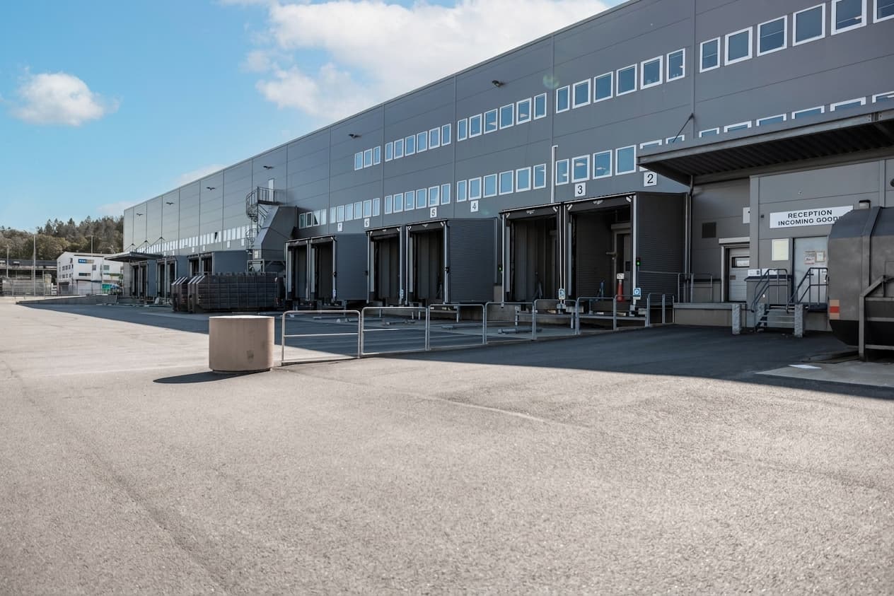 A large industrial warehouse with multiple loading bays, numbered doors, and a lorry parked near the reception area on a sunny day.
