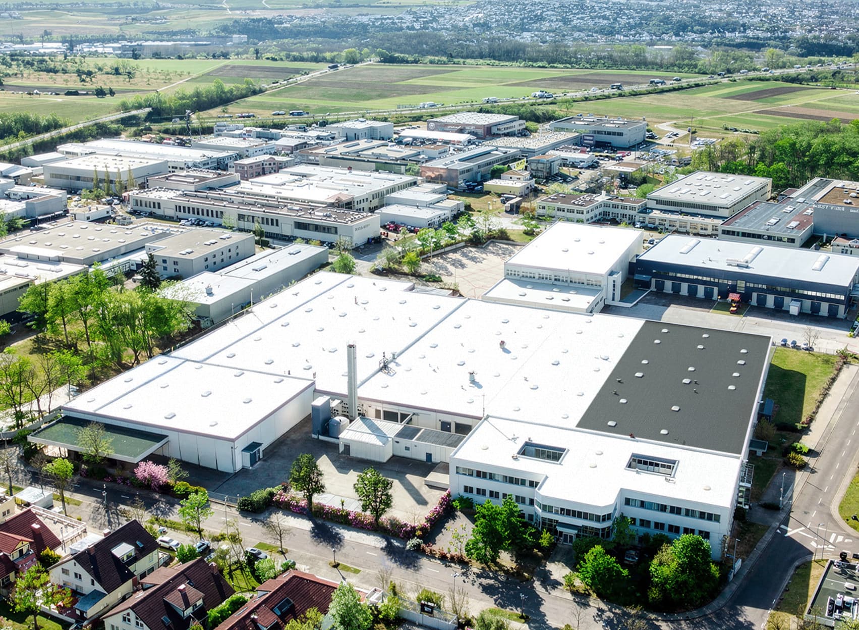 Aerial view of an industrial complex with large white-roofed buildings, surrounding roads, and green fields in the background.